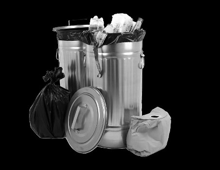 A large, cylindrical metal rubbish bin with a closed lid and two metal handles, positioned on a plain surface. The bin is filled to the brim with assorted waste, including paper and plastic items, some of which are spilling over the top. To the left of the bin is a tied black rubbish bag, leaning against it, and to the right is a crumpled white bag or paper wrapper resting on the ground. In front of the bin's lid, a smaller, flat container lies with its open side facing outward. The background is solid black, emphasizing the metallic finish and textures of the waste containers and bags, consistent with a waste collection or rubbish removal context in a commercial or residential setting, possibly in the UK, such as Wallington in the postcode area. The scene suggests an ongoing rubbish removal process, typical of services provided by Commercial Waste Wallington.