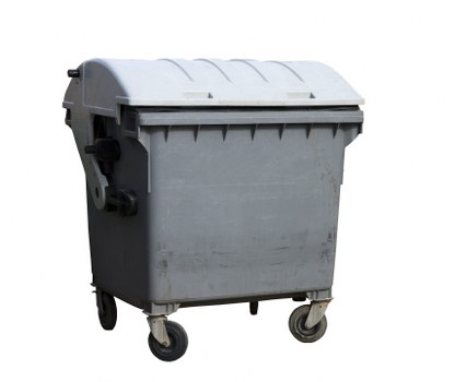 A large grey plastic wheelie bin with a hinged lid, positioned outdoors on a paved surface. The bin features a textured surface with vertical ridges along the sides and a black handle on the front for opening the lid. It is mounted on four small black wheels, with two visible at the front, allowing mobility for rubbish collection. The background includes a portion of a driveway or street, indicative of an urban or suburban environment, possibly in the vicinity of Wallington. The lighting appears natural, with subtle shadows cast by the bin, emphasizing its three-dimensional form. This image relates to waste management services such as rubbish collection and disposal, which involve companies like Commercial Waste Wallington, operating in local areas including SW postcode districts. Typically, such containers are used for general waste or commercial rubbish removal in residential or commercial premises within the town.