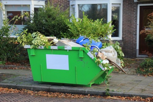 A large, bright green plastic skip situated on a paved driveway in front of a residential brick house with white-framed windows. The skip is filled with a mixture of green leafy branches, cardboard boxes, a white plastic bag, and a bundle of white materials, all piled to the brim and overflowing onto the sides. The surrounding area includes some lush green bushes and trees adjacent to the house, with a paved pathway and a small curb directly in front of the skip. The scene appears to be daytime with natural light illuminating the area, reflecting the typical rubbish removal activity handled by Commercial Waste Wallington in the local area near Wallington, Surrey, suggesting a domestic or small-scale clearance. The arrangement emphasizes the standard use of a skip for excluding miscellaneous waste, including garden greenery, packaging materials, and general household rubbish, consistent with professional waste management services in the region.